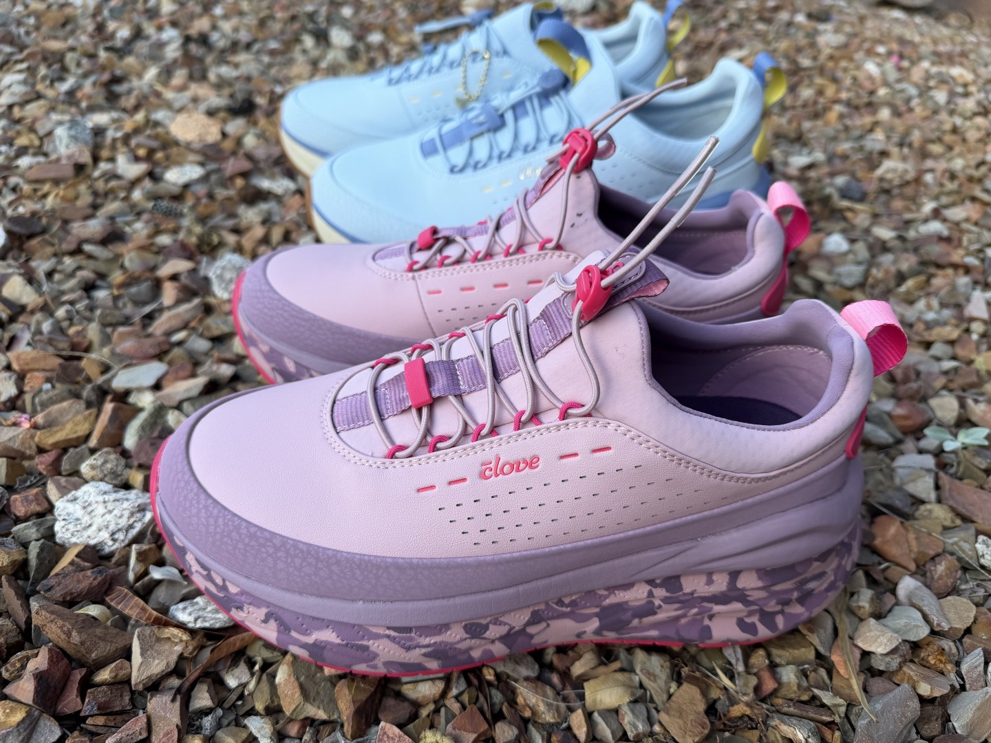 Two pairs of Clove Forte 2 on gravel — a mauve and pink women's pair in front and a light blue men's pair behind. Household two-tester.